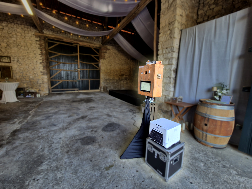 Rustic event space with stone walls, draped fabric, and string lights; a wooden photo booth stands next to a printer and equipment table.